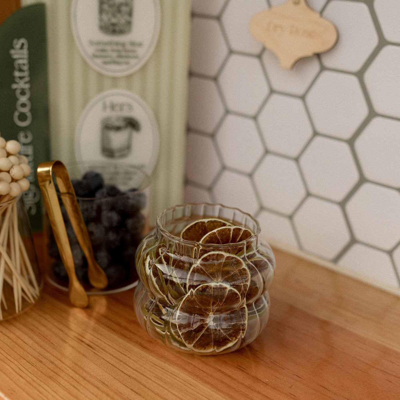 A close detail shot inside the Piaggio Tap Bar — brass cocktail tongs, a glass jar of dehydrated citrus wheels, blueberries in a crystal vessel, cocktail picks in a bud holder, all set on the solid wood bar top against the white hex-tile back wall with the 'Cocktails' menu cards.