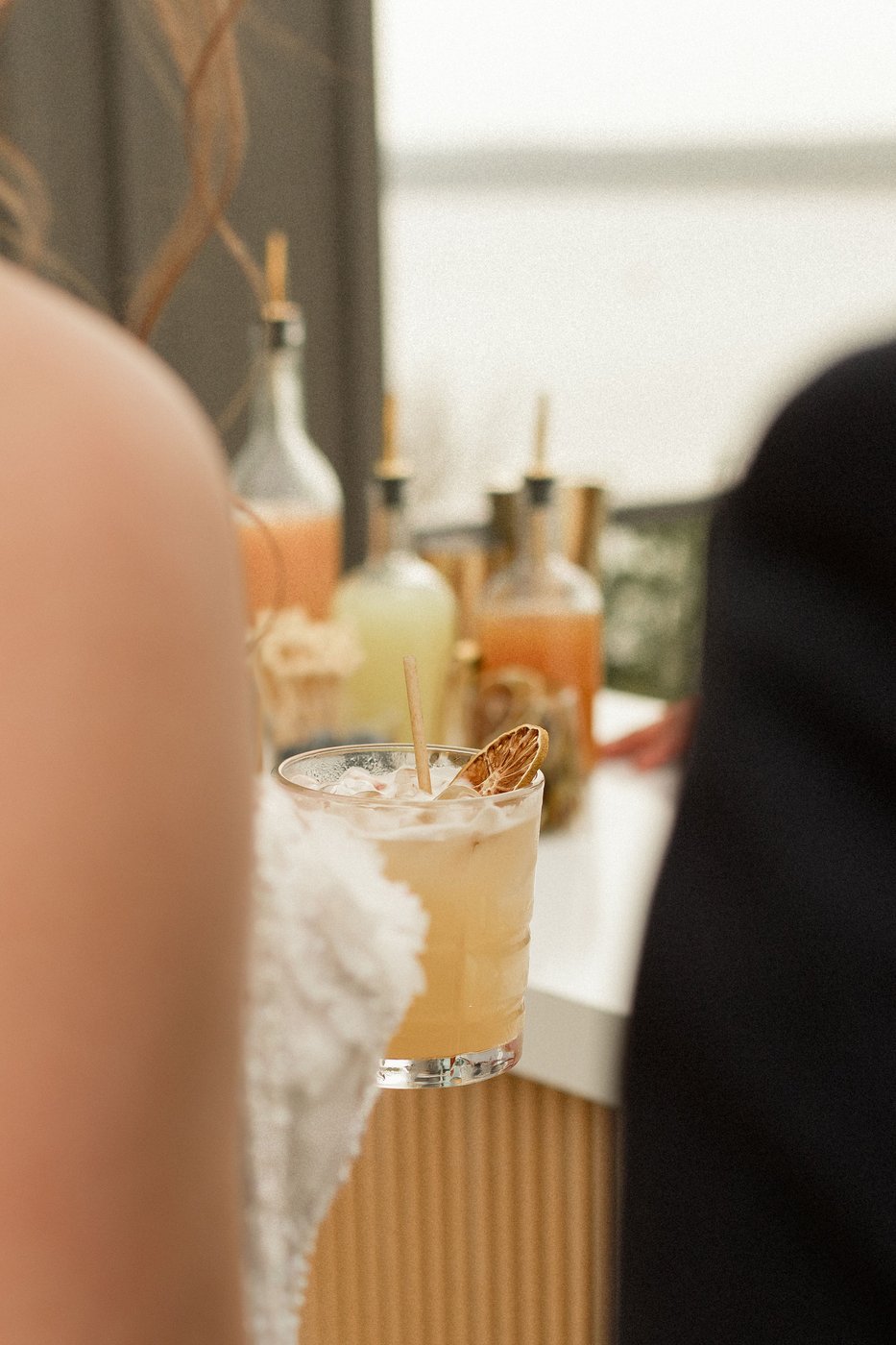 A close-up shot of a handcrafted cocktail with a dried lime garnish on a Satellite Bar Station, with Veronica and Augusto's silhouettes blurred in the foreground and Uphill Taps ingredient bottles in the background — the work happening behind the bar.