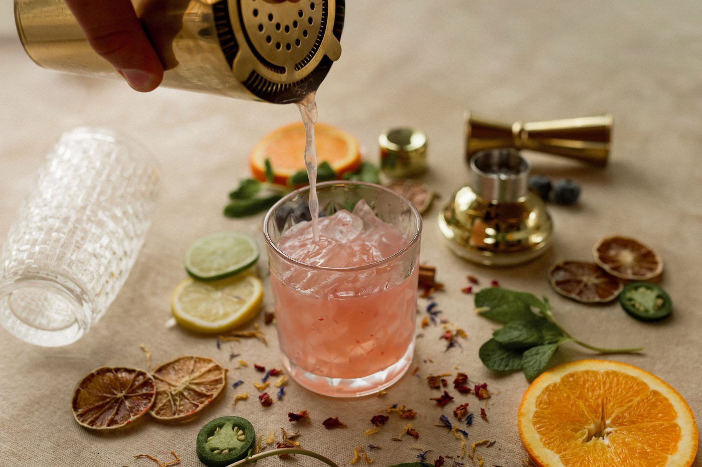 An Uphill Taps bartender pouring a cocktail from a brass shaker into a crystal glass, surrounded by fresh citrus, herbs, and dried botanicals — overhead view of the craft in motion.