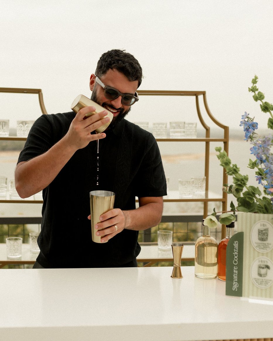 An Uphill Taps bartender pouring a cocktail mid-shake — brass shaker in hand, against a styled outdoor bar with florals and the Signature Cocktails menu card.