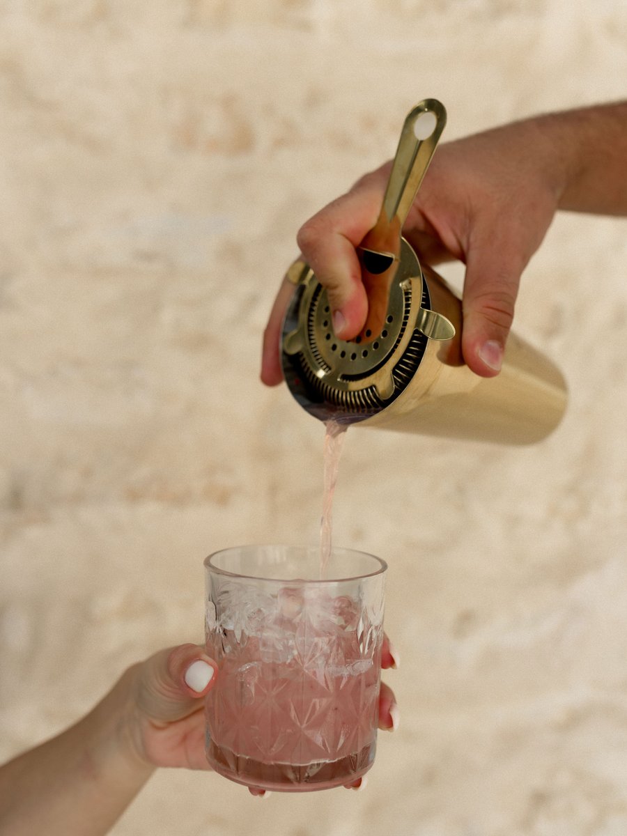 An Uphill Taps cocktail being poured from a brass shaker through a strainer into a crystal glass — close detail of the cocktail program in motion.