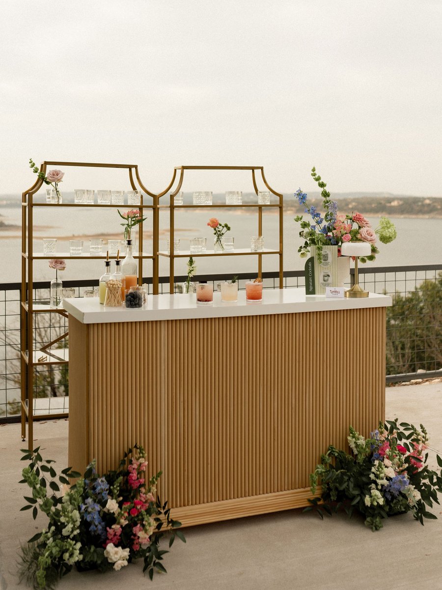 An Uphill Taps Satellite Bar Station — slatted wood-front bar with white quartz top, two gold-framed back walls, signature cocktails on the bar, styled florals at the base, with Lake Travis behind.