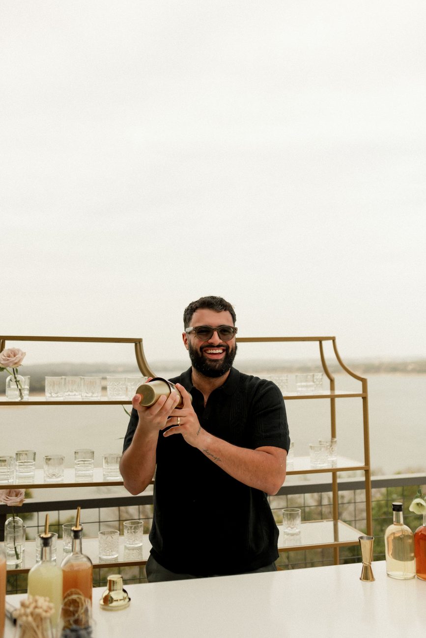 Augusto Fossi, co-founder of Uphill Taps, smiling and shaking a brass cocktail shaker behind a styled bar with Lake Travis behind him.