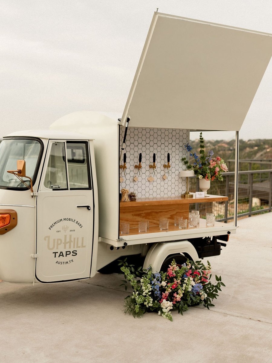 The Uphill Taps Piaggio Tap Bar — a vintage cream-colored Italian three-wheeler with cargo door open, branded UPHILL TAPS card on the side, hex-tile back wall with six brass taps, solid wood bar top, and styled florals at the wheel.