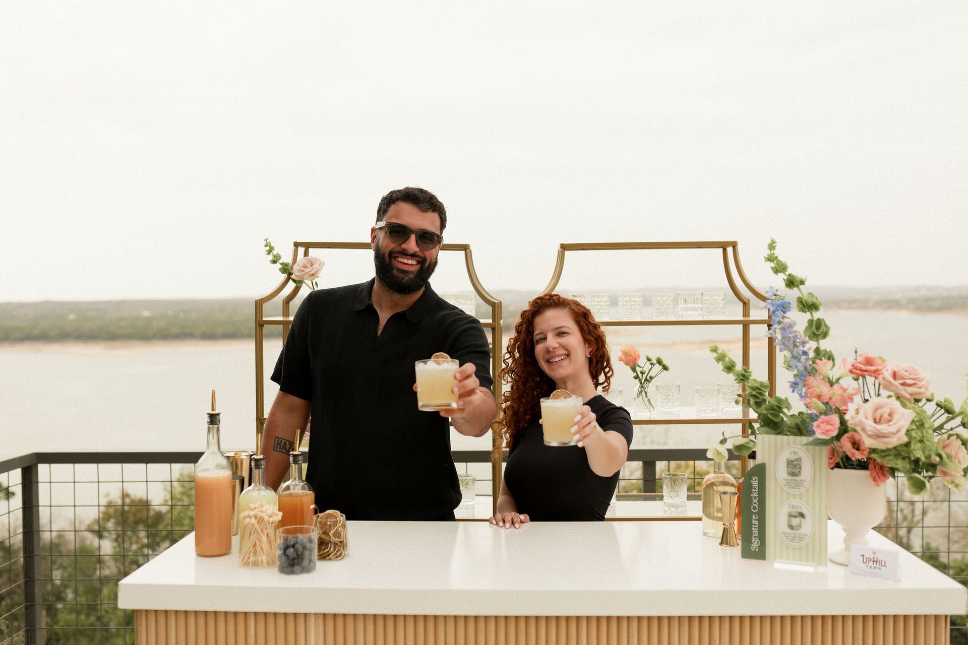 Uphill Taps founders Veronica and Augusto Fossi behind a styled white bar at a Hill Country overlook above Lake Travis, both holding handcrafted cocktails forward to camera and smiling, with gold-framed shelving of crystal glassware, pink roses and blue delphinium florals, and signature cocktail menu cards on the bar top.