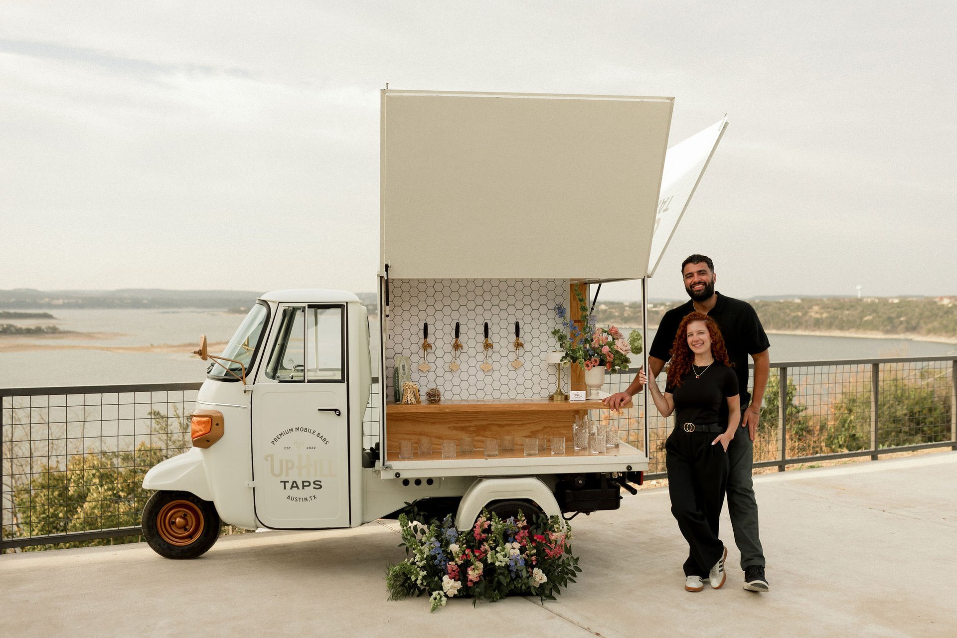 The Uphill Taps Piaggio mobile tap bar set up at a Hill Country overlook above Lake Travis, with founders Veronica and Augusto Fossi standing alongside, four taps poured, glassware lined along the bar top, and a floral arrangement at the base of the truck.