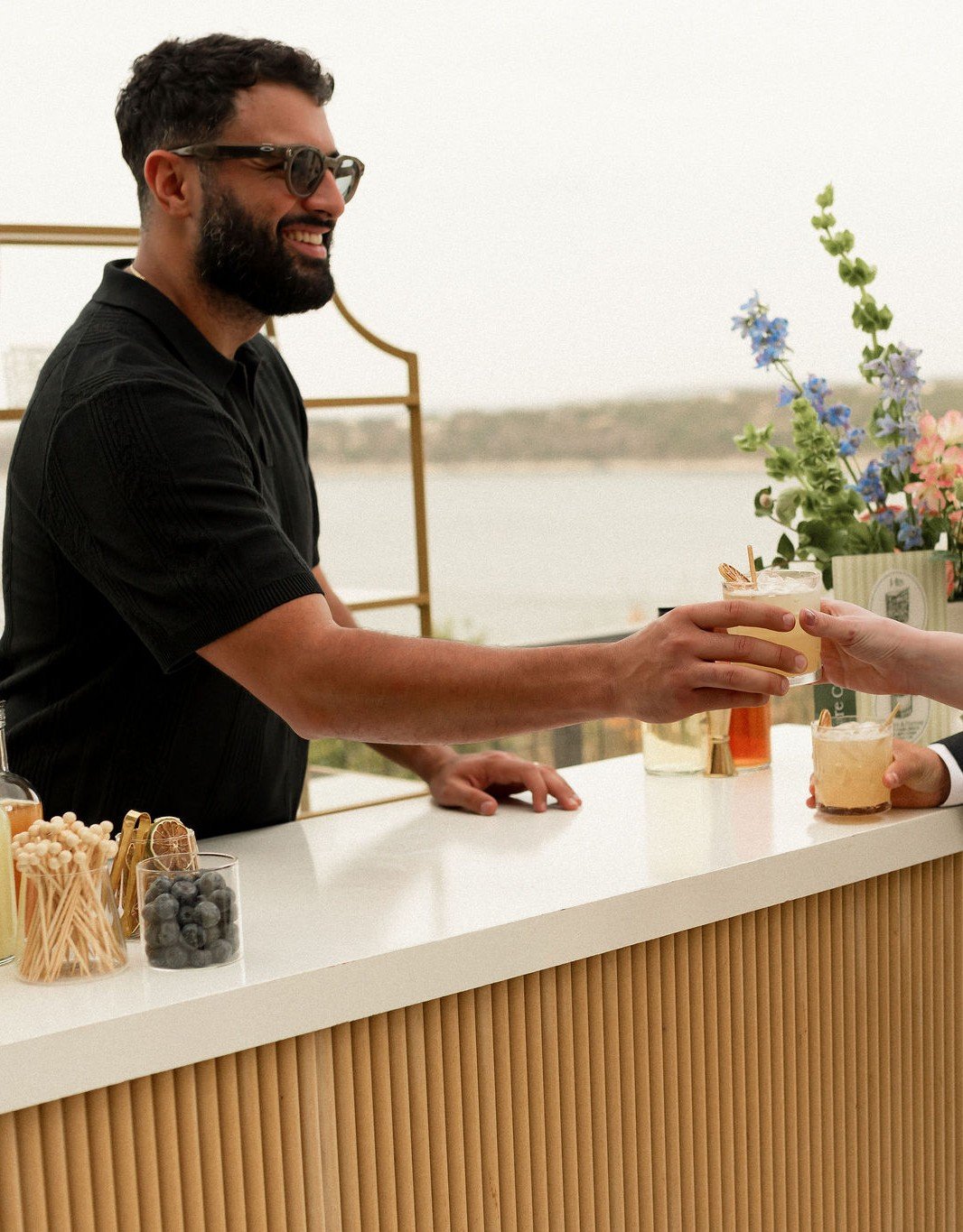 Augusto Fossi behind the Satellite Bar Station handing off a citrus-colored signature cocktail across the white marble bar top — wearing dark sunglasses and a black knit polo, smiling. The fluted cream-and-oak bar front, garnish jars with matchsticks and fresh blueberries, a brass jigger, and bottles of fresh juice are visible along the bar top. The gold-framed back wall, a styled floral arrangement of pink roses and blue delphinium, and the Lake Travis horizon are visible behind.