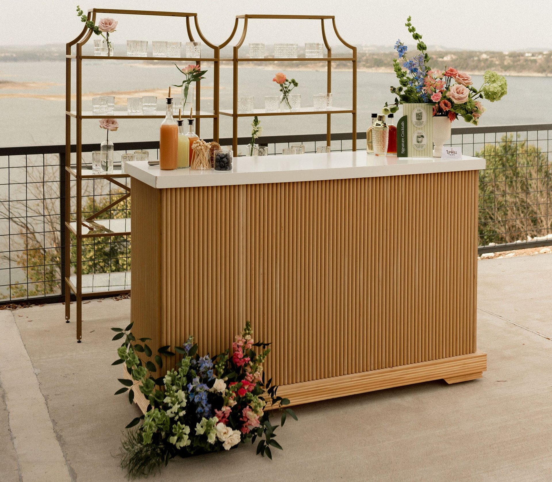 The Uphill Taps Satellite Bar Station set up at a Hill Country overlook above Lake Travis — a slatted wood-front bar with white marble top, twin gold-framed back wall with glass shelves and crystal glassware, pink rose and blue delphinium florals at the base, the Signature Cocktails menu card and UPHILL TAPS card on the bar top, and three signature cocktails plated on the bar.
