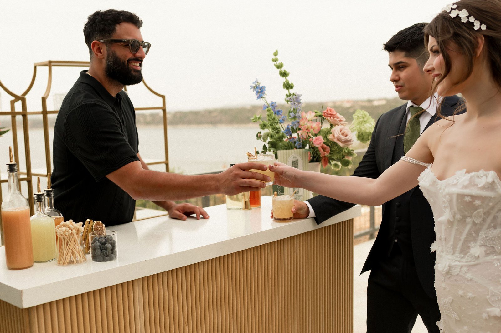 Augusto Fossi behind the Satellite Bar Station passing a fresh handcrafted cocktail across the white marble bar top to a bride and groom — the bride in a beaded white wedding dress, the groom in a charcoal suit with a green tie. Lake Travis, Hill Country horizon, and the gold-framed back wall with crystal glassware visible behind, with garnish jars, citrus, and bottles of fresh juice along the bar top in the foreground.