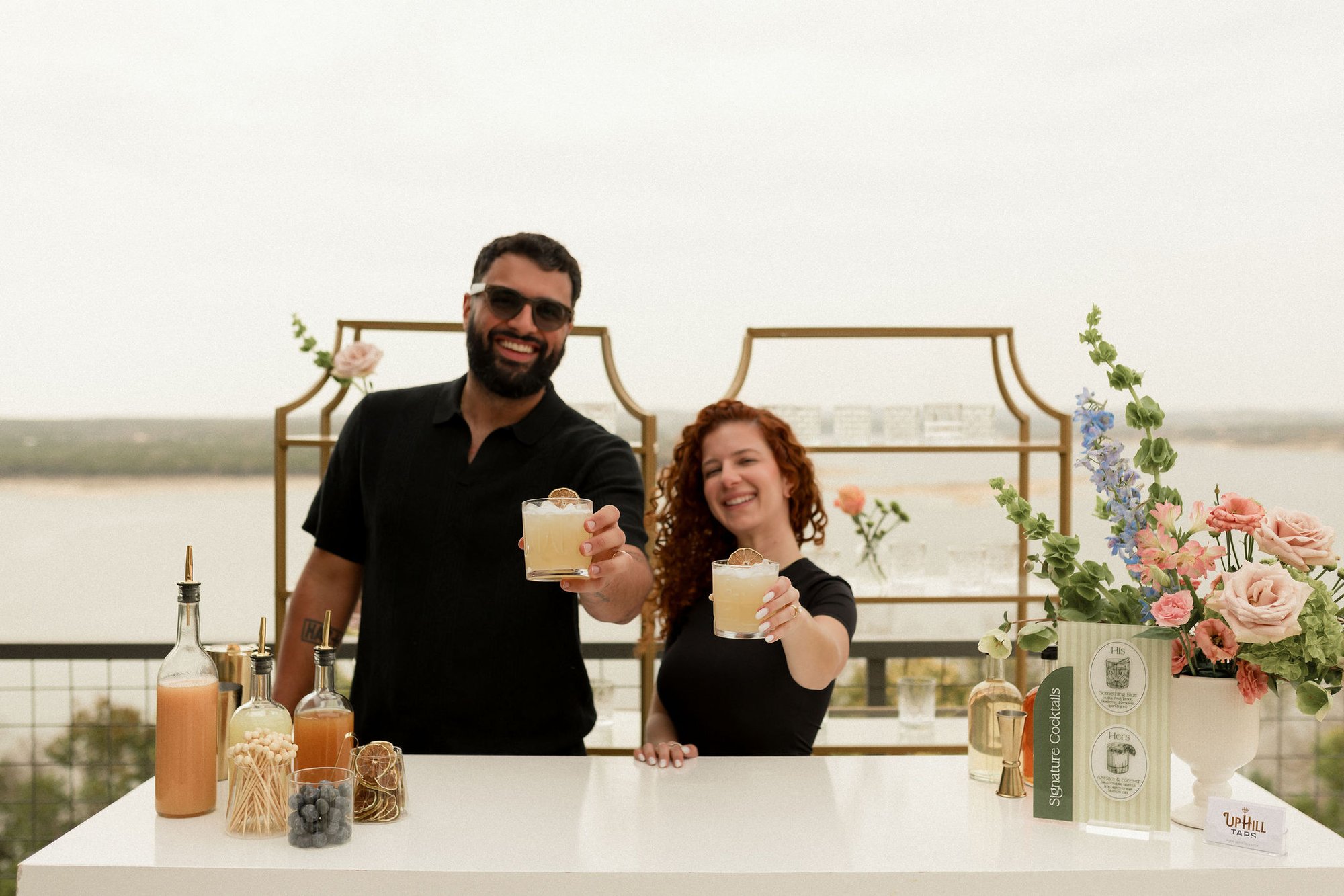 Veronica and Augusto Fossi, founders of Uphill Taps, behind the Satellite Bar Station holding handcrafted cocktails forward toward camera and smiling — with the gold-framed back wall, styled florals of pink roses and blue delphinium, signature cocktail menu cards, ingredient bottles on the bar, and Lake Travis and the Hill Country horizon as the backdrop.