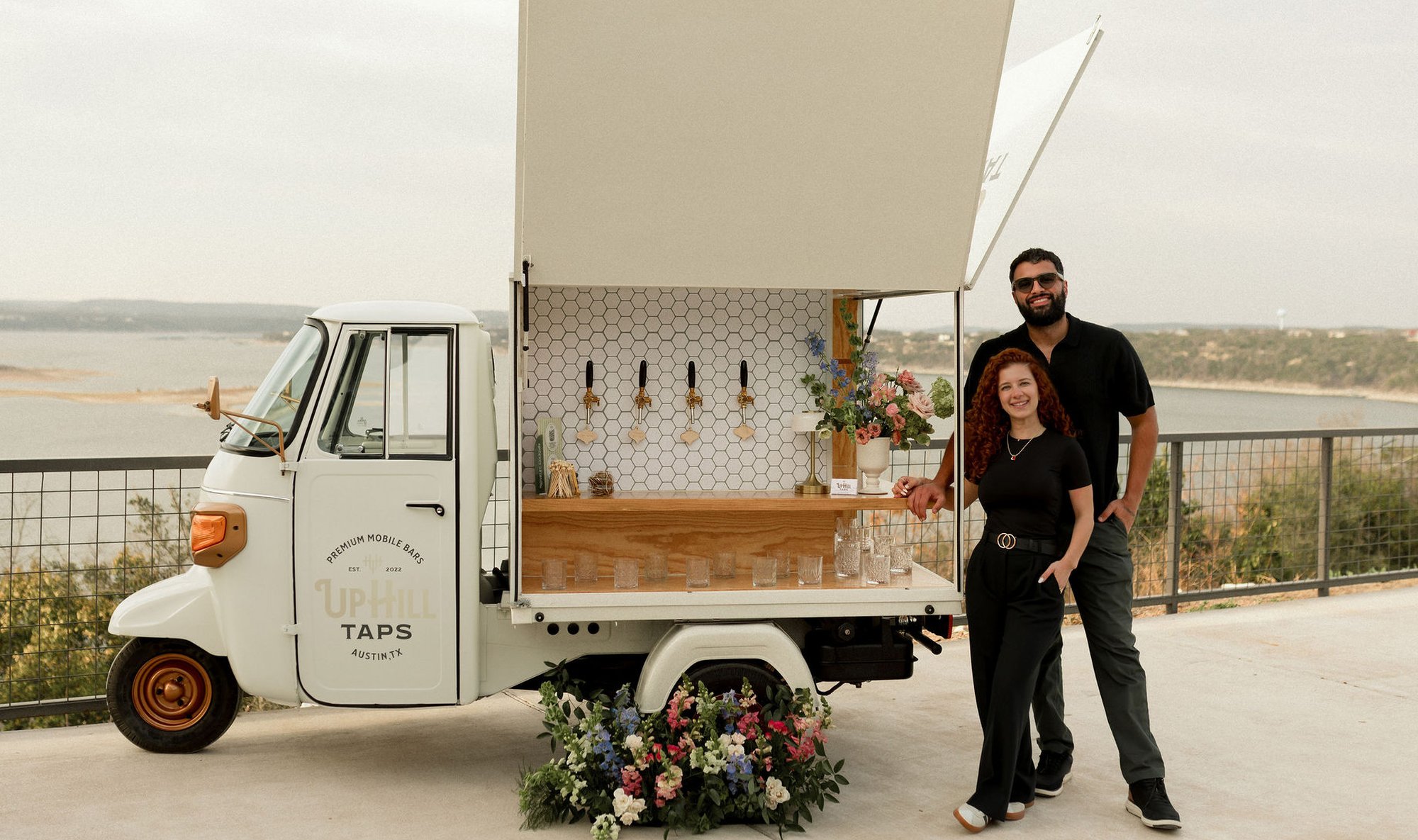 Veronica and Augusto Fossi standing beside the Uphill Taps Piaggio Tap Bar — a vintage cream-colored Italian three-wheeler with cargo door open, the 'UPHILL TAPS Premium Mobile Bars Est. 2022 Austin TX' branded card on the door, hex-tile back wall with four brass taps inside, wood bar top, styled florals at the wheel, and Lake Travis and the Hill Country horizon as the backdrop.
