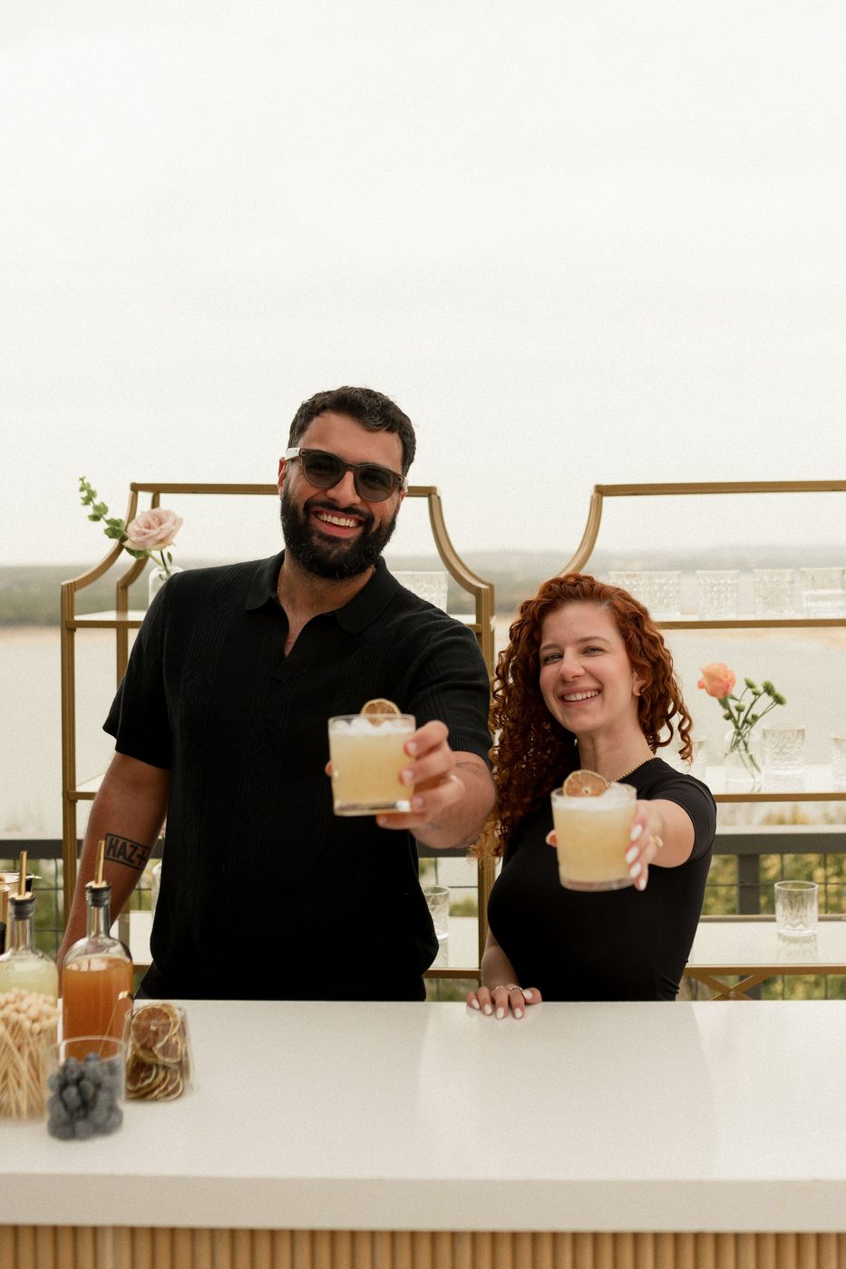 Veronica and Augusto, founders of Uphill Taps, behind a styled bar overlooking Lake Travis at golden hour, raising two cocktails with dried-citrus garnishes.